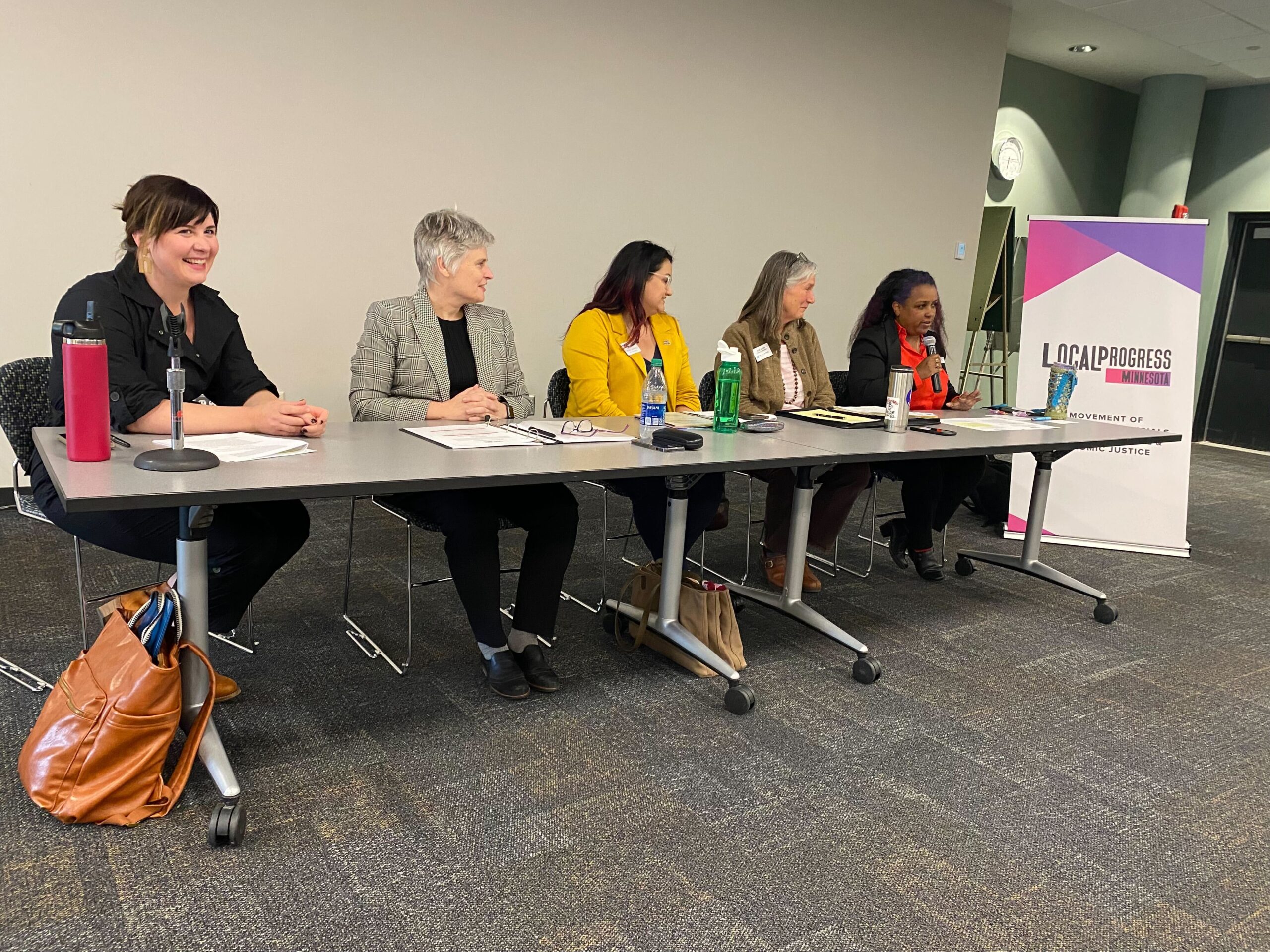 Four panelists (Mary Moriarty, Sue Budd, Kimberly Wilburn, and Heidi Garrido) and moderator Jillia Pessenda behind a table next to a Local Progress Minnesota sign.