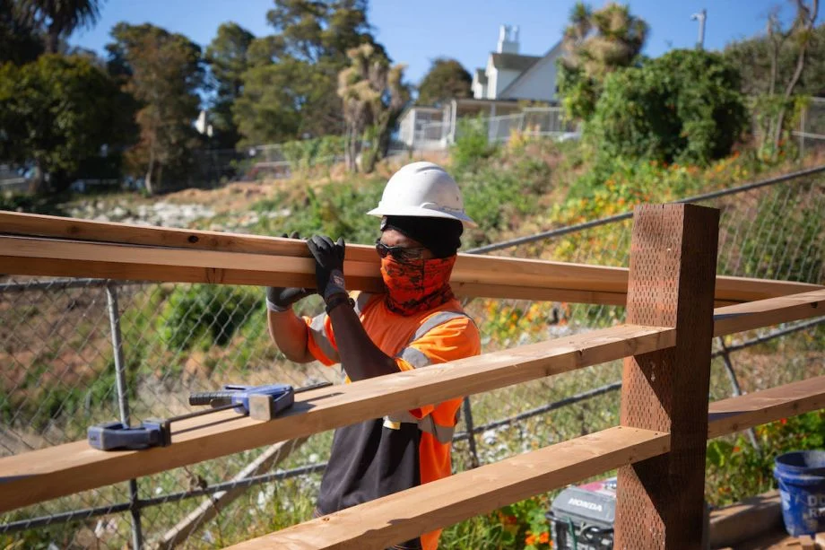 A construction worker carrying a large piece of wood in the hot sun with their face and body mostly covered up. (Photo courtesy Ivette Carolina Agudelo-Lopez + Yerba Buena Engineering & Construction, Inc)
